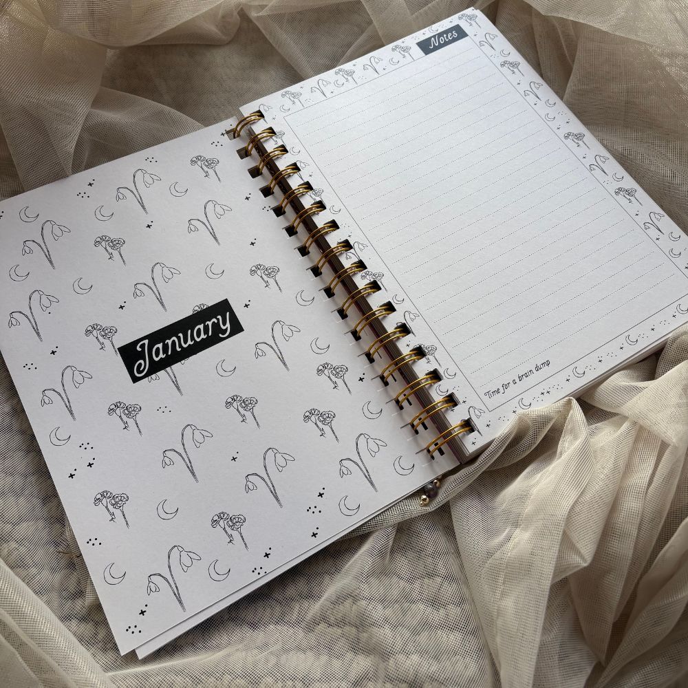 Open spiral-bound planner with decorative birth flower illustrations pages on a soft fabric background